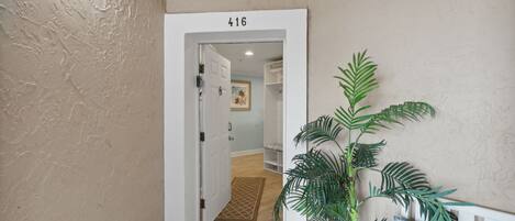Un pasillo con una puerta identificada como '416' que da a una habitación con piso de madera y un tapete. En primer plano, hay una planta en maceta y un banco con cojín.
