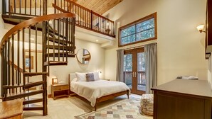 Une chambre à coucher avec un grand lit, un escalier en bois, un miroir rond, une table de chevet, une commode et une vue sur l’extérieur par une fenêtre.