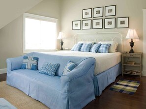 A bedroom with a bed, a blue sofa, a nightstand, a lamp, a window, and framed pictures on the wall.