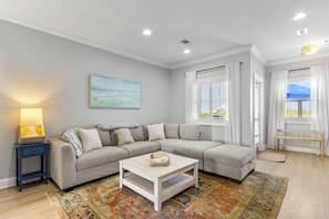A living room with a sectional sofa, a coffee table, a lamp, a bench, and a window with curtains. - Beautiful Town Home Beach Rental In Perdido Key Florida - Pool + Beach Access (Perdido Key)