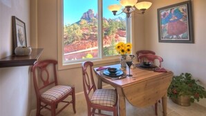 Un comedor con una mesa de madera, dos sillas rojas, un jarrón con girasoles y vistas a formaciones rocosas rojizas a través de un ventanal grande.