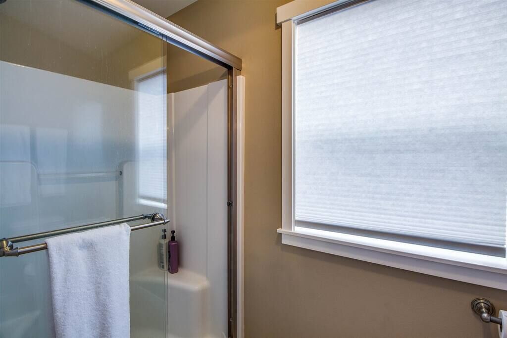 Une salle de bain avec une porte de douche en verre, un porte-serviettes avec une serviette blanche, une bouteille de shampoing et une fenêtre avec des stores.