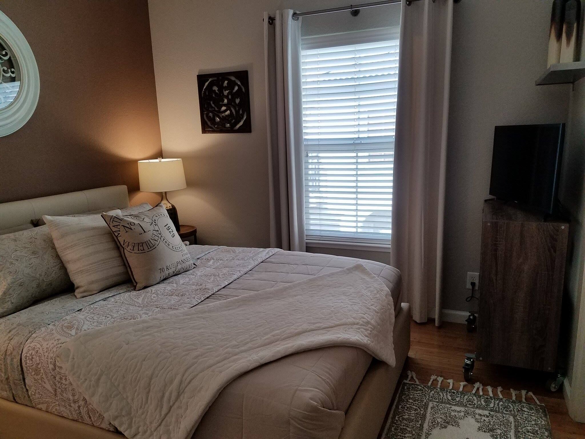 Uma cama bem arrumada com um edredom acolchoado e almofadas decorativas, uma luminária de cabeceira, uma janela com persianas e uma televisão sobre um suporte de madeira.