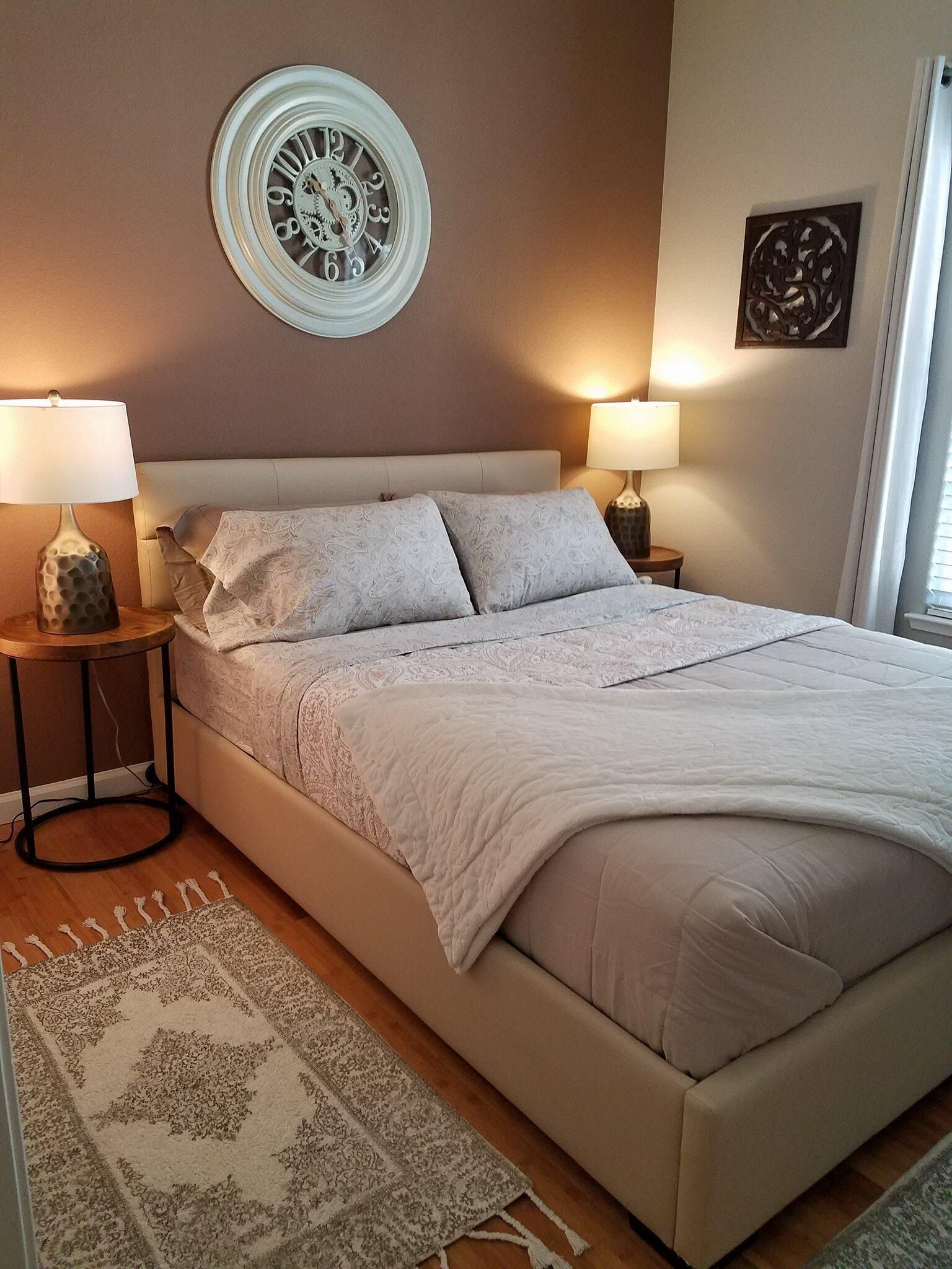 A neatly made bed with a white comforter and pillows, two bedside lamps, a decorative wall clock, and a patterned rug on a wooden floor.