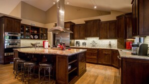 A spacious kitchen with dark wood cabinets, a large island with stools, stainless steel appliances, and a high ceiling with recessed lighting. - Hidden, Lush Treasure Close to Everything...Your Own Private Resort! (Mesa)