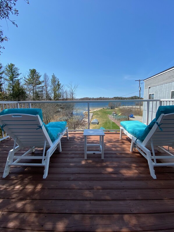 A wooden deck with two white lounge chairs and a small table, overlooking a lake and grassy area.