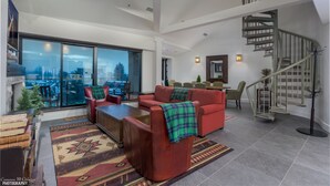 A spacious living room with a red sofa, a coffee table, and a large window offering a city view. A spiral staircase is visible in the background. - Top Floor. Top Views. Top of Whistler. 
The Penthouse at the Carleton Lodge. (Whistler)