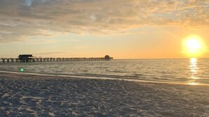 A beach with a pier extending into the water, a sunset in the background, and a sandy foreground with a pair of sandals. - Relax on the oversized lanai featuring an outdoor kitchen and mid-water view. (Naples)