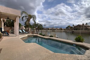 Piscina con vista a una masa de agua, rodeada de palmeras y un área de patio cubierta con sillones de descanso.