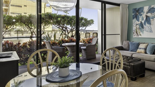 Ein modernes Wohnzimmer mit Glastisch, Korbstühlen, einer Couch und einem Balkon mit Pflanzen und Möbeln.
