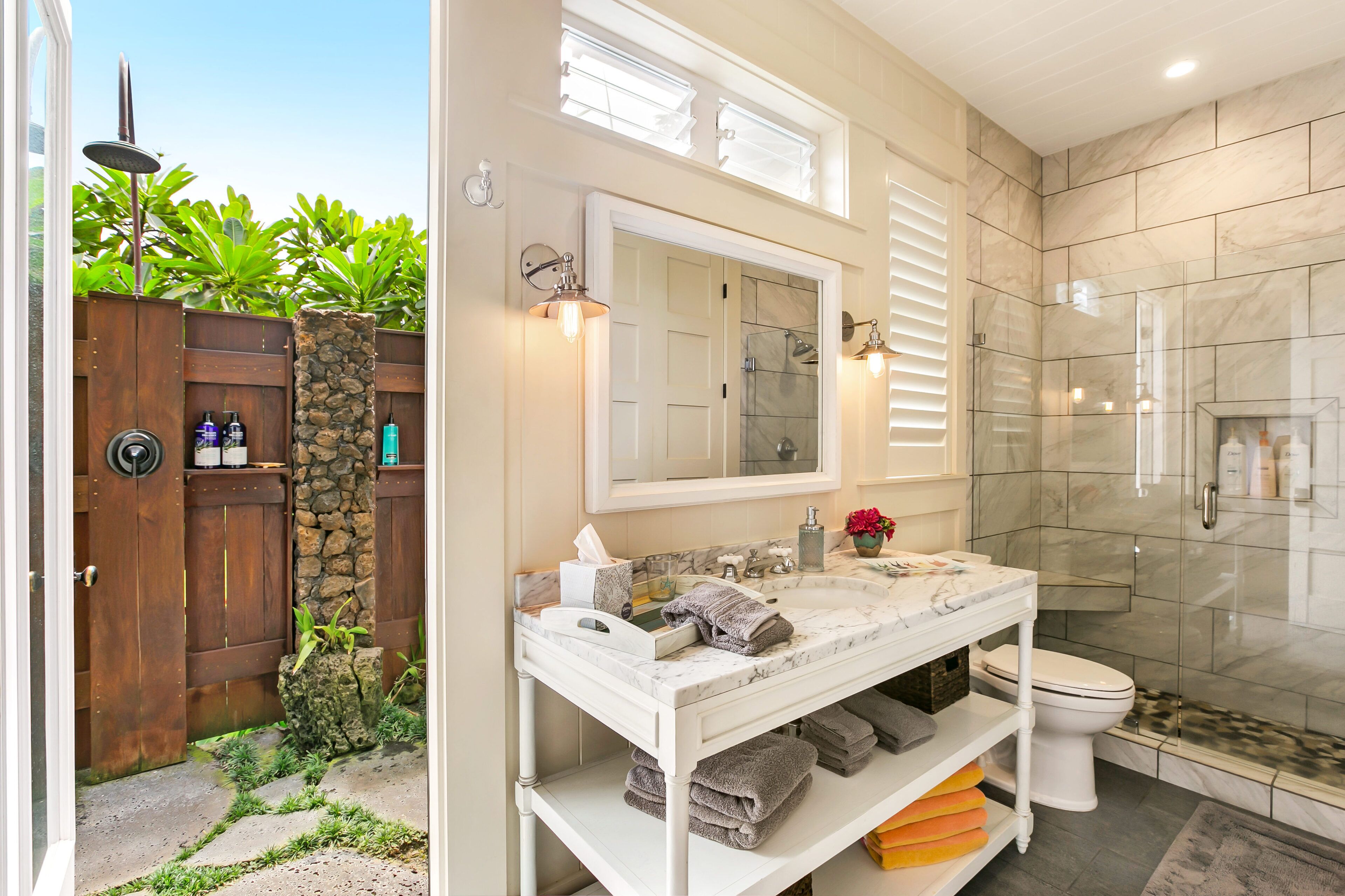 A bathroom with a marble sink, a large mirror, a glass shower enclosure, and a wooden door leading to a garden.