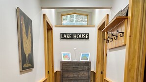 Un couloir aux murs lambrissés, une enseigne indiquant « MAISON AU BORD DU LAC », et une commode ornée de cadres contenant des photos.