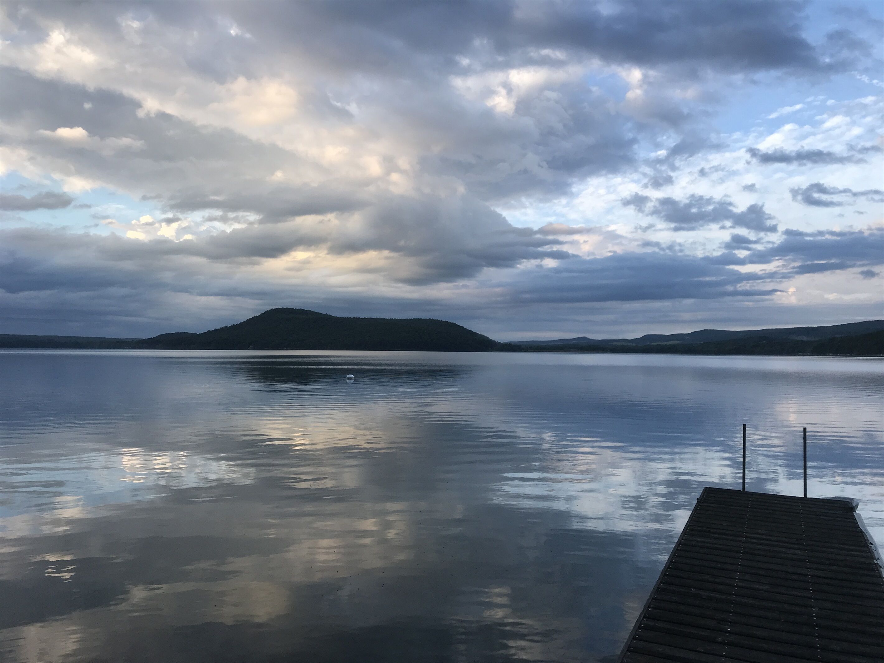 En stilla sjö med en träbrygga som sträcker sig ut i vattnet, omgiven av kullar och en molnig himmel.
