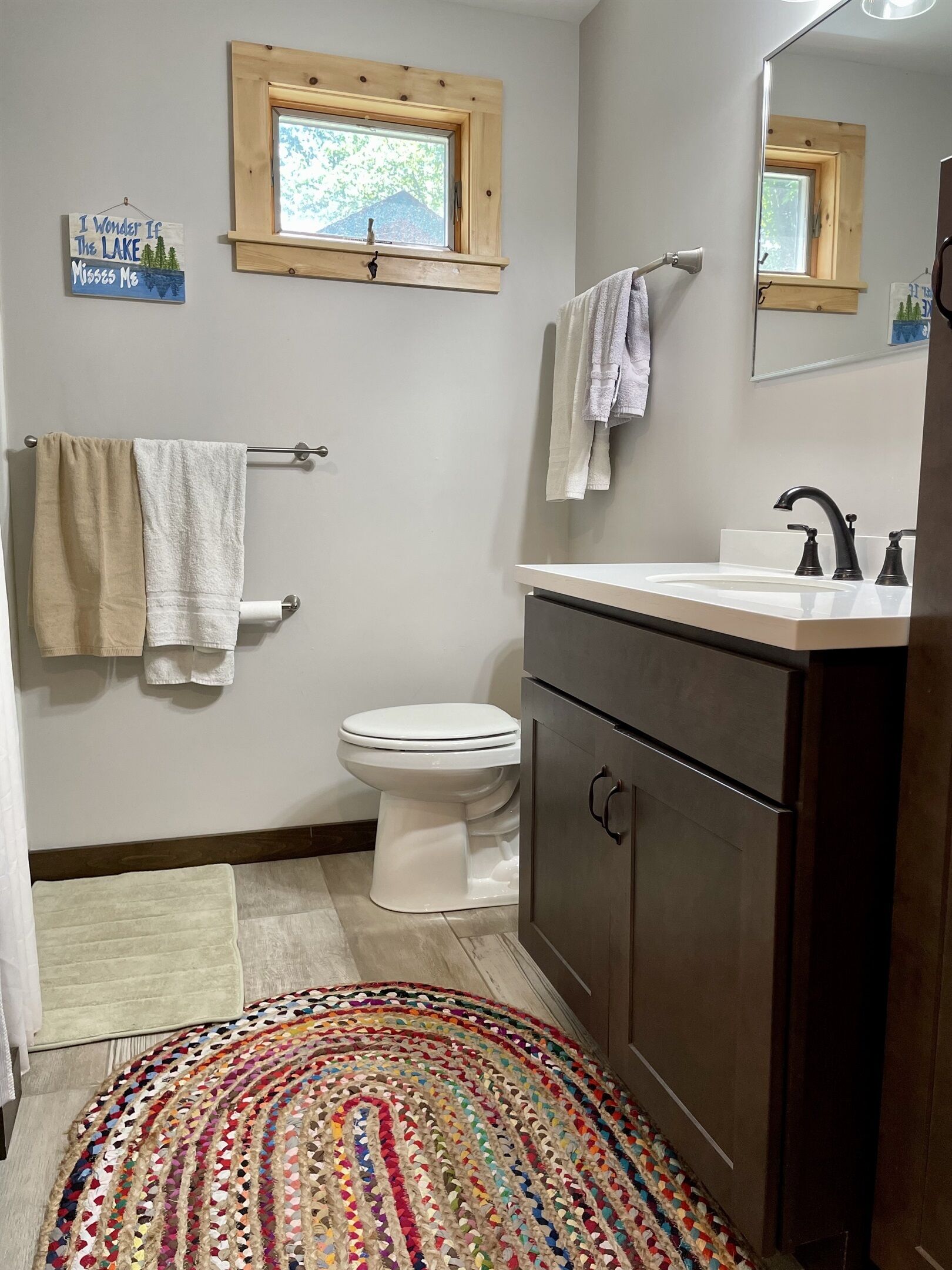 Un bagno con un WC bianco, un lavandino con rubinetto scuro, una finestra in legno e un tappeto colorato tessuto.