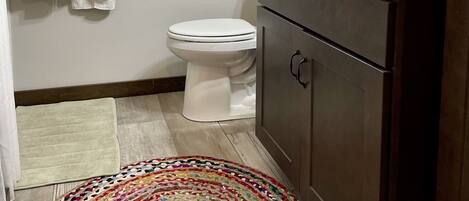Un bagno con un WC bianco, un lavandino con rubinetto scuro, una finestra in legno e un tappeto colorato tessuto.