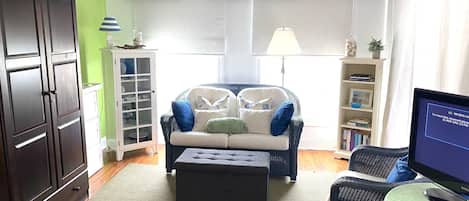 Een woonkamer met een donker houten kast, een rieten bank met kussens, een kleine tafel met een afstandsbediening en een boekenkast.