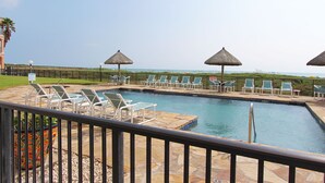 A pool area with lounge chairs and thatched umbrellas, surrounded by a stone deck and a fence. - Experience comfort in this 2-bedroom condo with a bonus room, perfect for 8 guests. (South Padre Island)