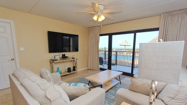 A hotel room with a sofa, a coffee table, a television, and a balcony with a pool view. - Experience comfort in this 2-bedroom condo with a bonus room, perfect for 8 guests. (South Padre Island)