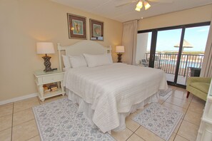 Habitación de hotel con una cama grande, dos mesitas de noche con lámparas, una silla, un balcón con vista al mar y una pequeña mesa con libros.