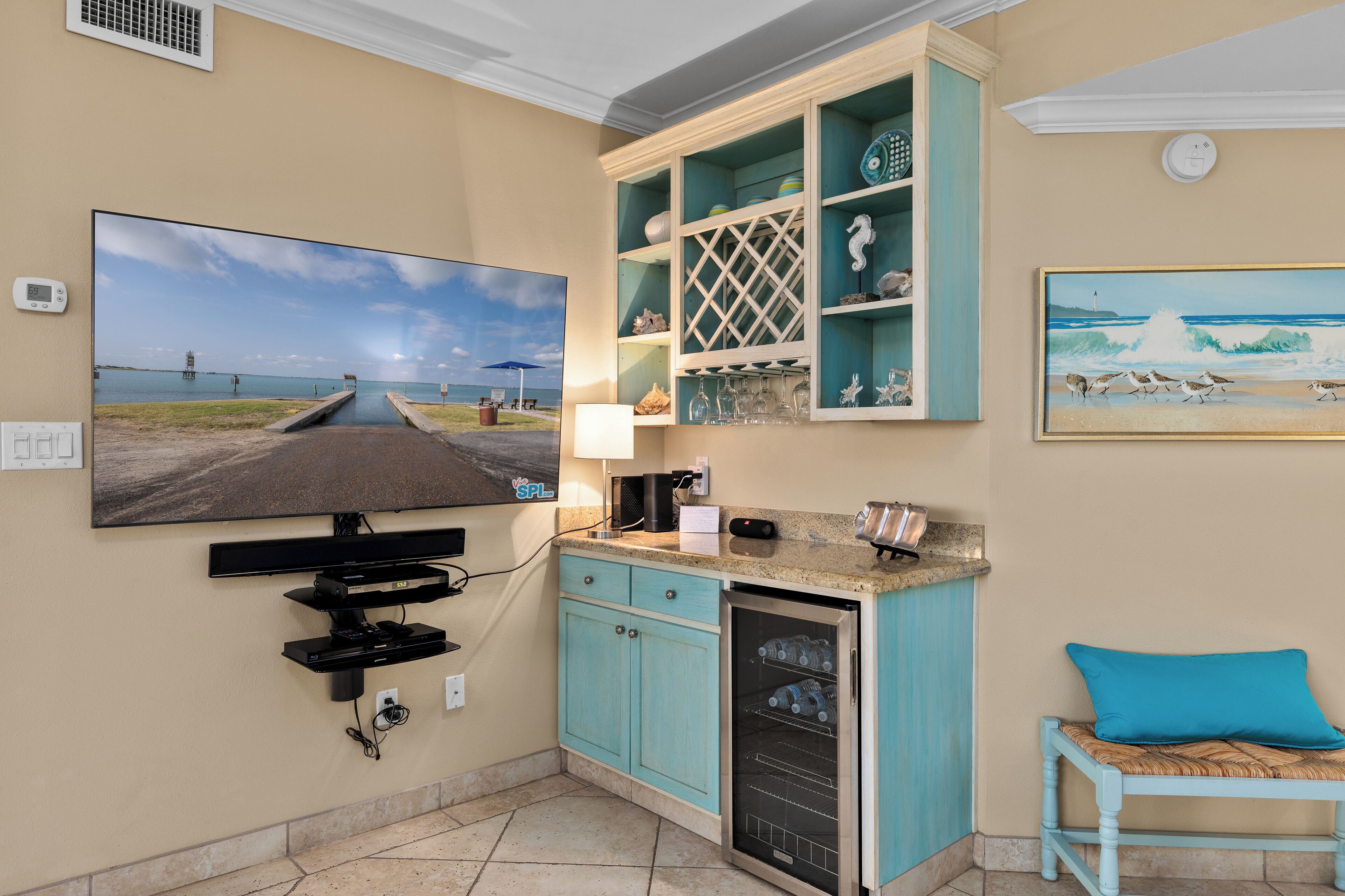 A kitchen with a large flat-screen TV mounted on the wall, a built-in refrigerator, and a teal-colored cabinet with glassware.