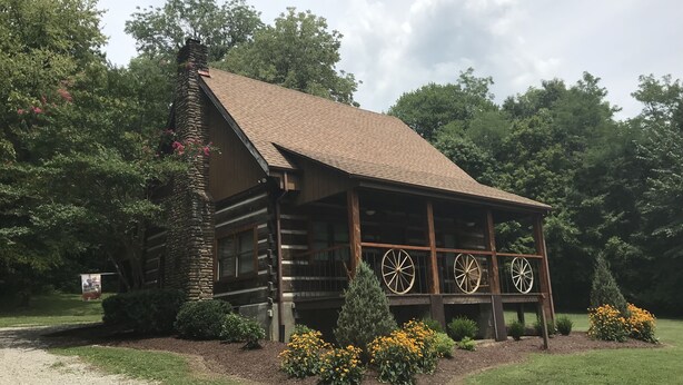 A rustic log cabin with a stone chimney, wooden wheel spindles, and a well-maintained garden with yellow flowers and shrubs. - The Cabin at Flat Rock Farms - rustic charm on a beautiful former horse farm  (Lewisburg)