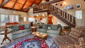 A spacious living room with a high ceiling, wooden beams, a staircase, and a kitchen area.