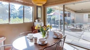 Un comedor con una mesa redonda, sillas, un jarrón con flores y vistas al exterior a través de ventanas amplias.