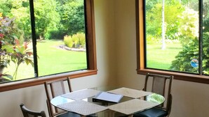 A corner room with a glass table and four chairs, two windows with a view of a garden.