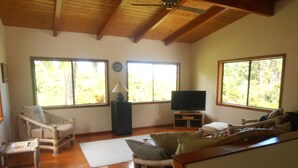A spacious living area with wooden floors, a ceiling fan, a television on a stand, and large windows offering a view of the outdoors.