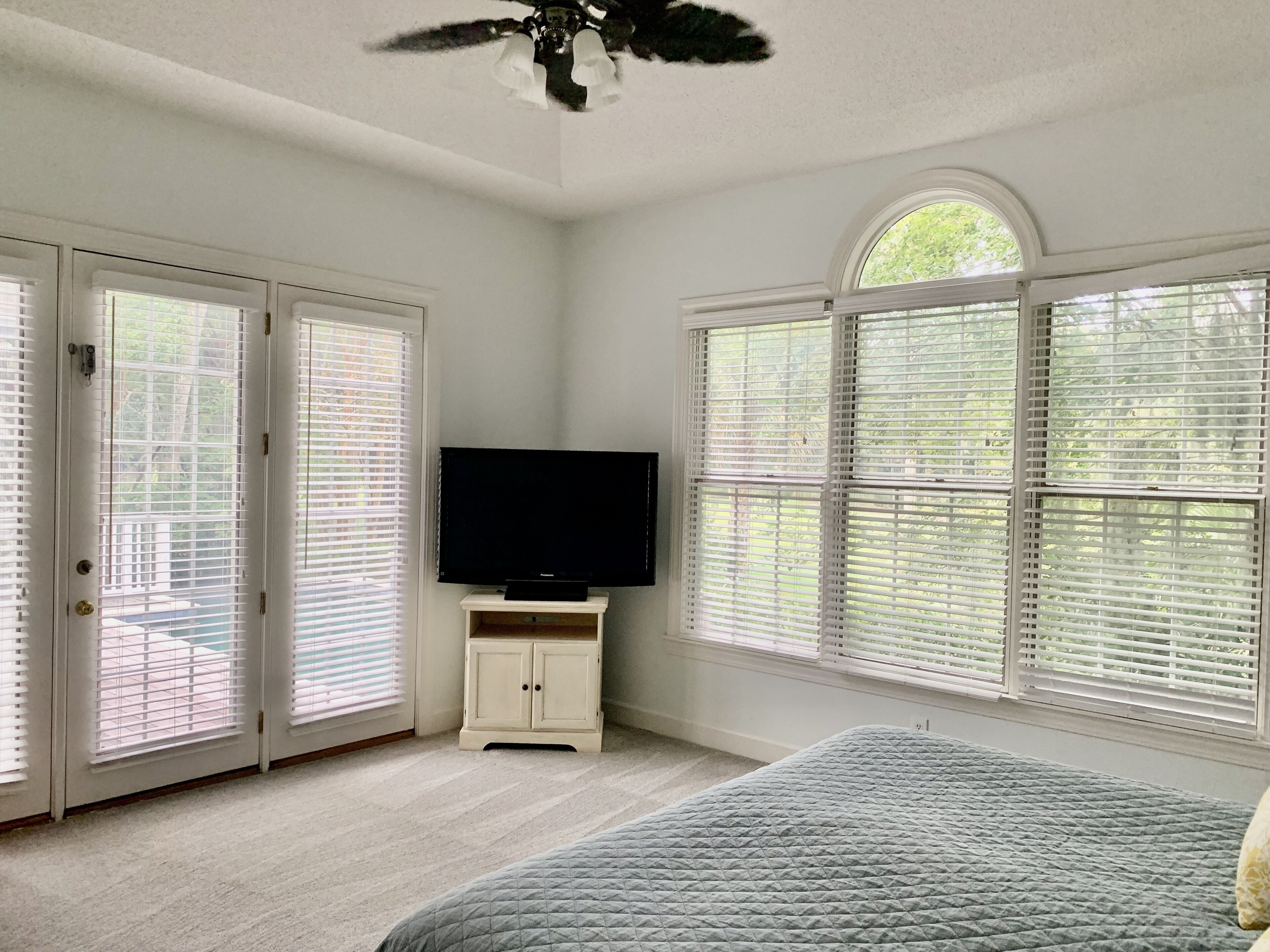 A bedroom with a bed, a television on a stand, a ceiling fan, and large windows with blinds.