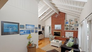 A spacious living room with a high ceiling, wooden beams, a brick accent wall, and a cozy seating area with a leather couch and a coffee table. - Crown Point Beach House - Beach and Water on 3 Sides! (San Diego)