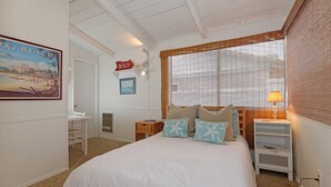 A bedroom with a bed, a nightstand, a window with blinds, a painting, and a sign on the wall. - Crown Point Beach House - Beach and Water on 3 Sides! (San Diego)