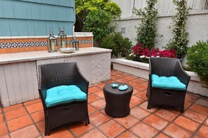 Dos sillas de mimbre con cojines color turquesa y una pequeña mesa redonda con posavasos color turquesa, sobre un patio con baldosas y un resguardo de azulejos, con plantas en macetas.