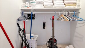 Een wasruimte met een plank vol opgevouwen handdoeken, een wasmand en verschillende schoonmaakspullen.