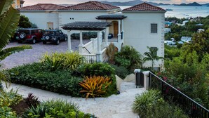 A two-story house with a covered patio, parked cars, and a view of the sea. - Guavaberry Villa, a St. John Villa with Three King Suites Near Cruz Bay (Cruz Bay)
