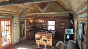 A rustic cabin interior with a wooden ceiling, a kitchen area with a stove and sink, two bar stools, and a couch.