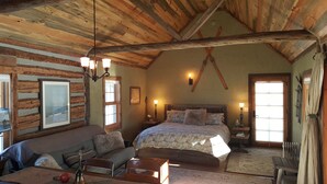 A rustic room with a wooden ceiling, a bed with patterned bedding, a wooden bench, a table with a vase, and a framed picture on the wall.