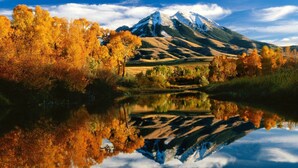 A mountain with a snow-capped peak, surrounded by autumn-colored trees and a calm lake reflecting the scenery.