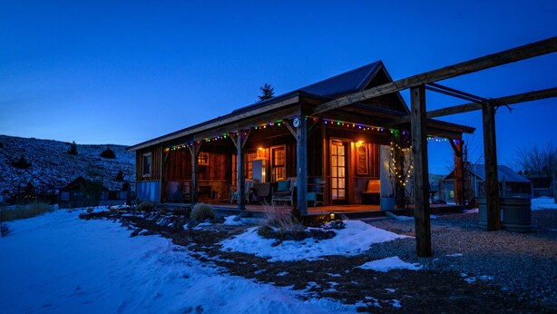 A wooden cabin with a porch, decorated with string lights, surrounded by snow.
