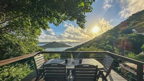 Une terrasse en bois, agrémentée d’une table et de chaises, offre une vue panoramique sur une baie où se trouvent des bateaux, le tout étant entouré d’une végétation luxuriante et de collines.