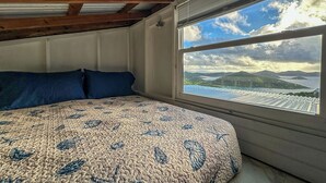 Une chambre avec un grand lit, une fenêtre donnant sur un paysage de montagnes et d’eau, et des poutres en bois au plafond.