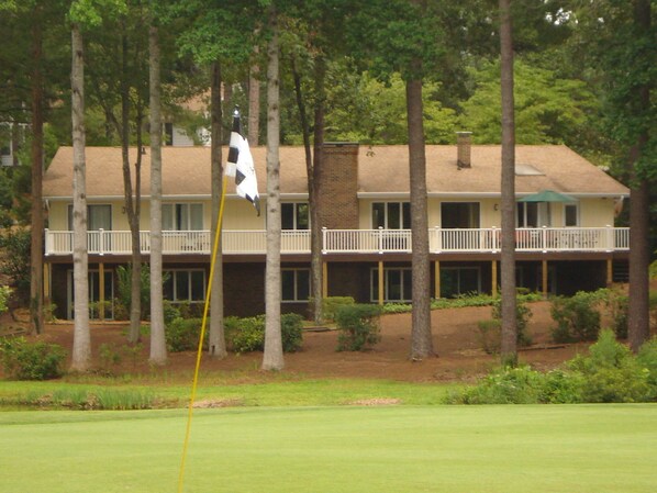 A golf course with a flagstick in the foreground, surrounded by trees. In the background, there is a two-story building with a balcony and a green umbrella.