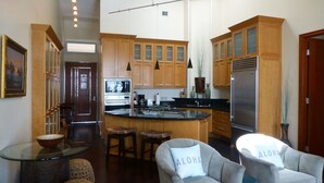 Cozinha moderna com armários de madeira, uma ilha central e eletrodomésticos de aço inoxidável. Há dois bancos para bar e uma poltrona cinza com almofada.