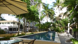Uma área de piscina com mesa e cadeiras de vidro, cercada por palmeiras e uma grade.