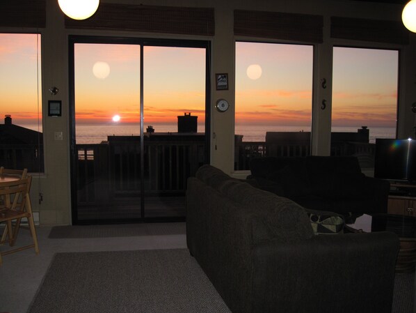 Una sala de estar con un gran ventanal corredizo que da a un balcón con vista al océano al atardecer.