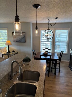 Cocina con encimeras de granito, lavamanos de acero inoxidable e iluminación colgante. Cuenta con un comedor con mesa y sillas de madera, y una ventana con persianas.