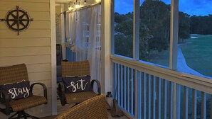 A covered outdoor seating area with wicker chairs and a table, a compass wall decoration, and a view of a golf course at dusk. - Beautiful Southport, Golf Frontage & Oak Island Beach club .New Quartz counters  (Southport)