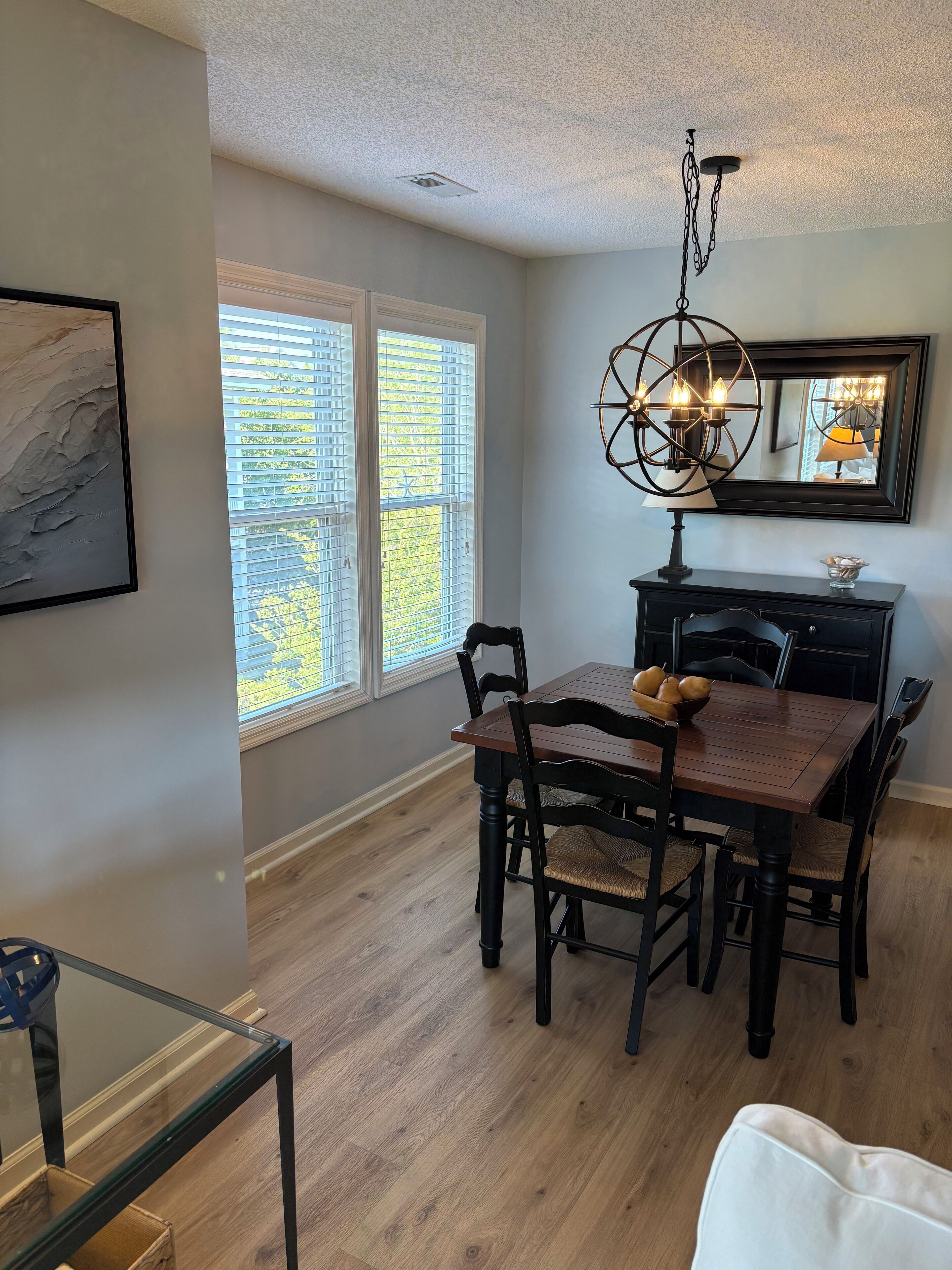 A dining area with a wooden table and chairs, a chandelier, and a large window with blinds.