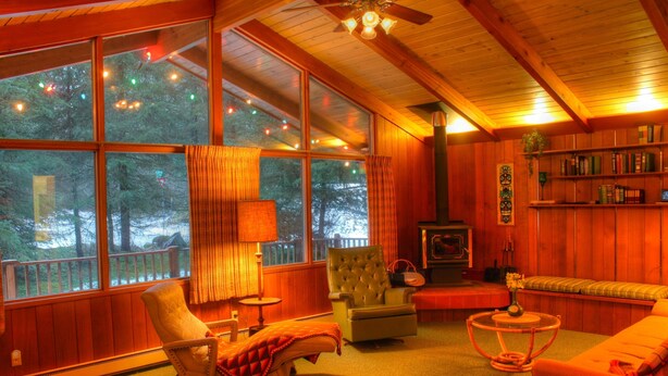 Interior acogedor de una cabaña con ventanas amplias, chimenea, estantería y una zona de estar cómoda.
