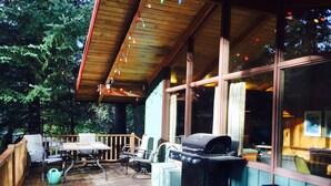 Una terraza cubierta al aire libre con una mesa y sillas de madera, una parrilla y vistas del bosque circundante.